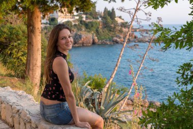 Güzel genç bir kadın oturuyor ve kameraya gülümsüyor. Arkasında deniz var.