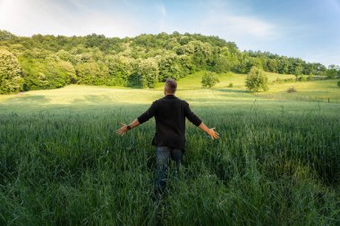 Genç adam buğday tarlasını kollarıyla kucaklıyor ve güzel manzaranın tadını çıkarıyor.