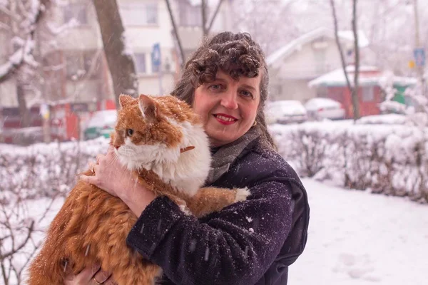 Kıvırcık saçlı, gülümseyen ve karlı bir günde turuncu bir kediyi tutan bir kadın.