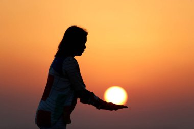 Deniz kıyısında duran, turuncu gökyüzünde güneşi tutan genç bir kadının silueti.. 