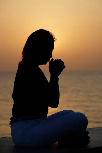 Silhouette at sunset of faithful woman praying, hands folded in worship to god. Concept for religion, faith, prayer and spirituality.