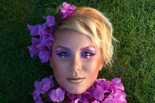 Mulher bonita deitada na grama, flores perto do rosto, maquiagem brilhante — Fotografia de Stock