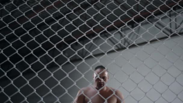 Svalnatý stíhač MMA se blíží k osmiúhelníkové kleci a dívá se do kamery. Bojovník se drží plotu v kleci.. — Stock video