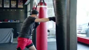 Caucasian handsome man wearing boxing gloves punching ahead on sandbag in gym or fitness club.