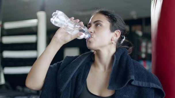 Fit woman in sportswear relaxing in gym and drinking water, equipment on background. — Video