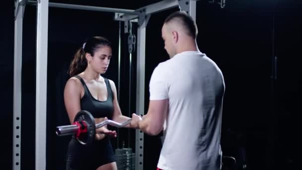 Молодой тренер, смотрящий на мускулистую женщину, делающую упражнения со штангой в тренажерном зале — стоковое видео
