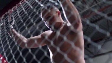 Muscular MMA fighter is standing by the octagon cage and looking down tired. Fighter is holding on to the fencing of the cage.