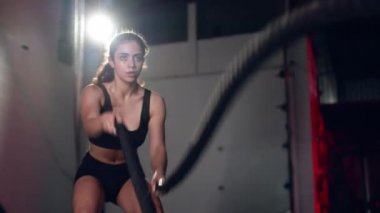 Active dedicated motivated female athlete training in gym with battle ropes.