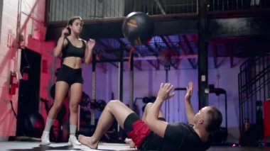 young athletic woman working out on abs with professional male coach, throwing cross fit ball at modern gym.