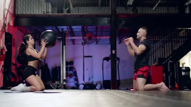 Mujer atlética joven haciendo ejercicio en abdominales con entrenador masculino profesional, lanzando la bola de cross fit en el gimnasio moderno. — Vídeo de stock