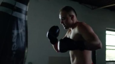 Caucasian handsome man wearing boxing gloves punching ahead on sandbag in the dark room.