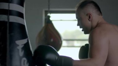 Caucasian handsome man wearing boxing gloves punching ahead on sandbag in the dark room and then leaning on the punching bag tired.