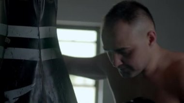 Caucasian handsome man wearing boxing gloves punching ahead on sandbag in the dark room and then leaning on the punching bag tired.