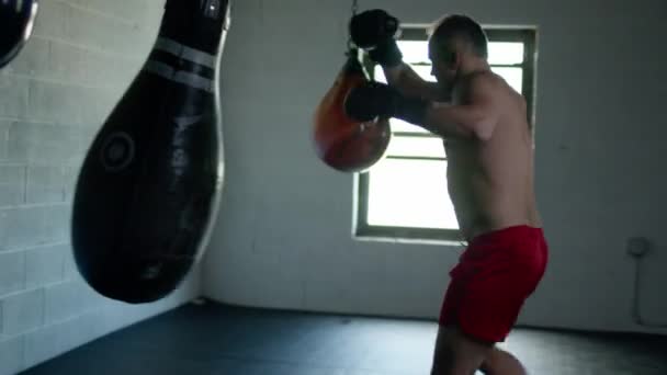 Kaukasischer gutaussehender Mann mit Boxhandschuhen schlägt im dunklen Raum auf Sandsack ein. — Stockvideo