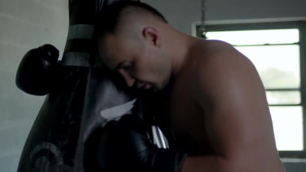 Kaukasischer gutaussehender Mann mit Boxhandschuhen, der im dunklen Raum auf einem Sandsack vorprescht und sich dann müde auf den Boxsack stützt. — Stockvideo