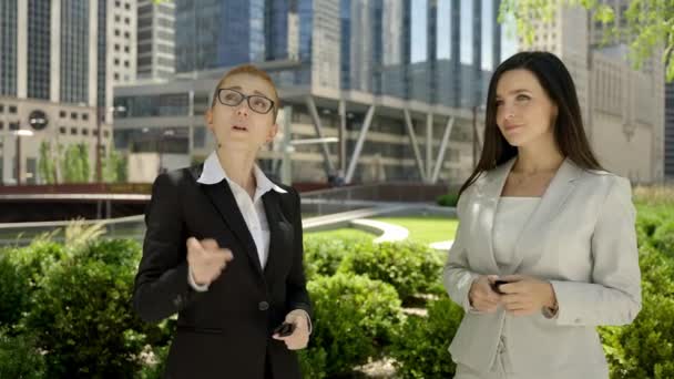 Two stylish cute women in beautiful business suits are standing in the city park and talking to each other. — Video