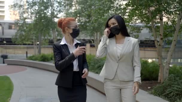 Two young Caucasian female busness women in face masks walking and talking in the park, taking face masks off and talking. — Video