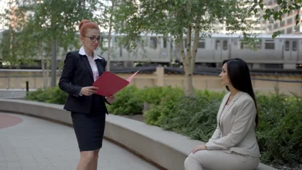 Twee stijlvolle schattige vrouwen in mooie zakenpakken zitten in het stadspark en praten met elkaar. — Stockvideo