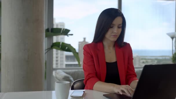 Mujer de negocios de confianza está sentado en la oficina moderna junto a la ventana panorámica con vista a la ciudad y trabajando en un ordenador portátil. — Vídeos de Stock