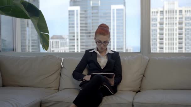 Belle jeune femme avec les cheveux roux assis sur le canapé dans la chambre et à l'aide d'un appareil tablette. — Video