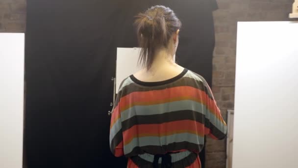 Chica artista con pinturas y pinceles pasea por el taller hasta el caballete. — Vídeo de stock