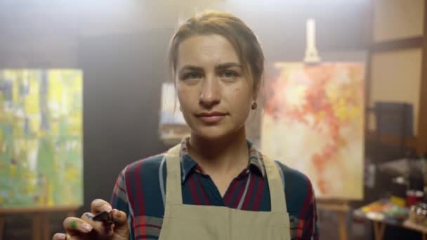 Femme artiste regardant avec confiance dans la caméra avec un pinceau et une palette dans ses mains. Séquence Vidéo