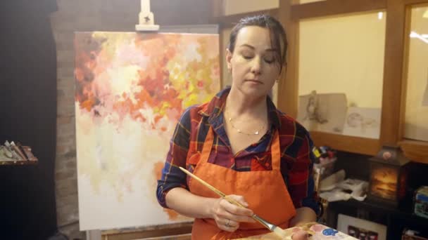 Femme artiste est debout près du chevalet et à la recherche d'inspiration avec un pinceau et une palette dans ses mains. — Video