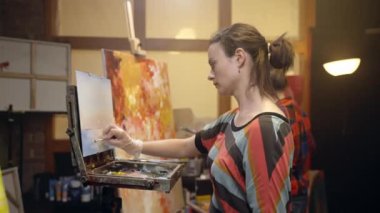 Kadın sanatçılar palet bıçağı ve yağlı boyayla tuvale resim çiziyorlar. Tuvale manzara çiziyorum. Sanatçı sehpaya yaklaşıyor..