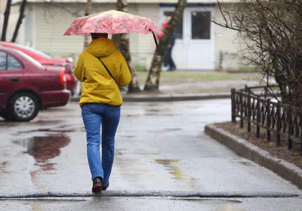 A young woman in blue jeans and a yellow jacket with a colored umbrella in the rain on the street, ulitsa Podvoyskogo, St. Petersburg, Russia, April 2021