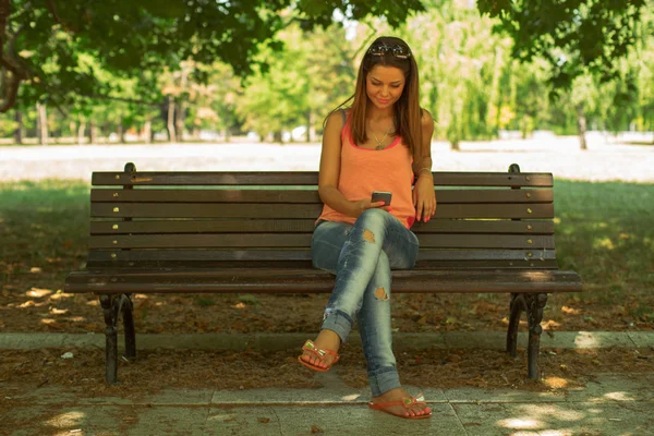 Mulher sentada no banco no parque e mensagens de texto no smartphone — Fotografia de Stock