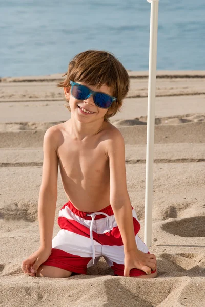 dizlerinin üzerinde güneş gözlüğü ile plaj şemsiyesi altında çocuk