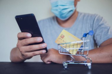Tıbbi maskeli bir adam evden online alışveriş için akıllı telefon kullanıyor.