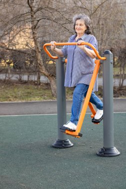 Dışarıdaki simülatörde spor yapan yaşlı bir kadın. Gri saçlı yaşlı kadın parkta egzersiz yapıyor.