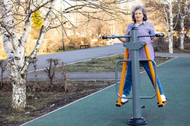 Dışarıdaki simülatörde spor yapan yaşlı bir kadın. Gri saçlı yaşlı kadın parkta egzersiz yapıyor.