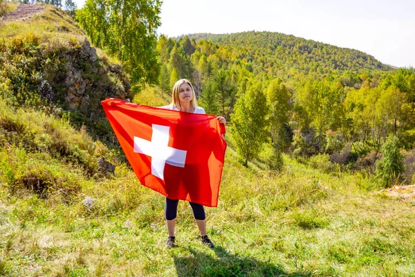 Egy európai megjelenésű nő tartja a svájci zászlót a fák és hegyek hátterében napsütéses időben. Szőke lány svájci zászlóval a kezében — Stock Fotó