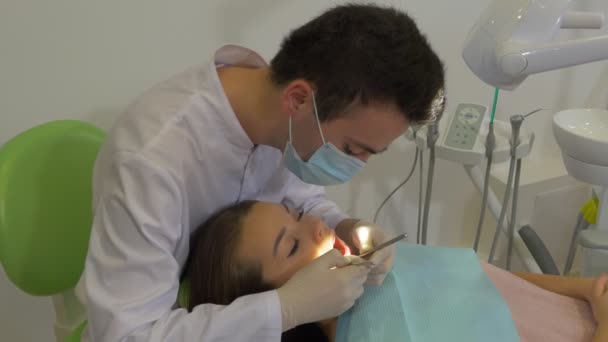 Dantista masculino na máscara está examinando um dentes Jovem Mulher Paciente Médico está usando um espelho bucal Olhando atentamente levanta uma lâmpada na sala de tratamento dentário — Vídeo de Stock