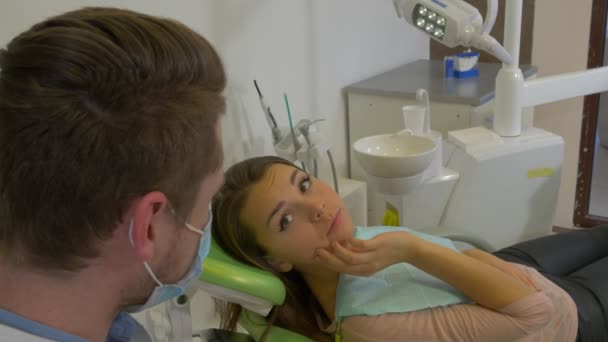 Chica se queja de un dolor de muelas tocando una mandíbula Hablar con un hombre dantista en máscara es escuchar a un paciente asiente examinando un diente en la sala de tratamiento dental — Vídeos de Stock