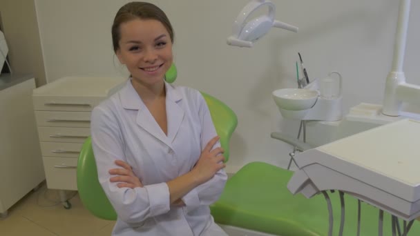Dantist в лабораторії пальто сидить її руки схрещеними і посміхаючись дівчина на зеленому кріслі в стоматологічній обробці кімната молода жінка дивиться на камеру щасливий лікар — стокове відео