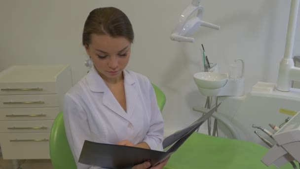 Dantist czyta notatki w Fold podnosi głowa dziewczyna w Lab płaszcz jest uśmiechnięty siedzi na krześle w Dental leczenie Pokój młody ładne Doktor Kobieta — Wideo stockowe