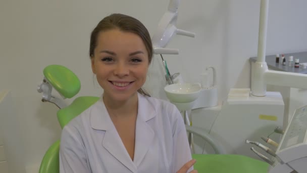 Dantista en abrigo de laboratorio sonríe sentada en una silla en la sala de tratamiento dental Mujer joven está sentada con los brazos cruzados Panorama de una silla verde — Vídeo de stock