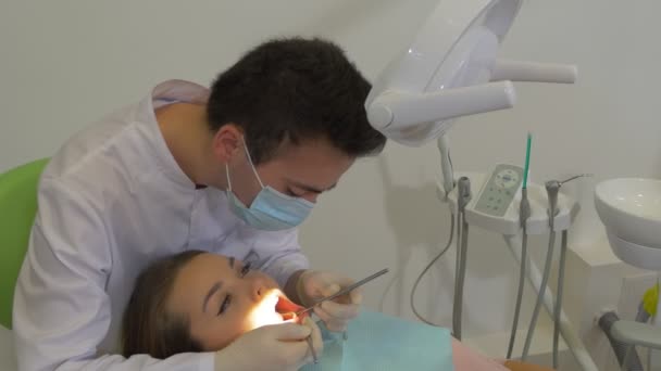 Dentista en máscara está sentado sobre un paciente examinando los dientes de un paciente por un espejo bucal en la sala de tratamiento dental Sillas de paciente verdes Cátedra del médico — Vídeo de stock