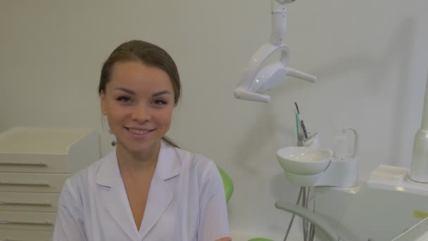 Dantista en abrigo de laboratorio está sonriendo sentado en una silla verde en la sala de tratamiento dental Mujer joven está mirando el Panorama de la cámara de una sala de hospital Ward — Vídeos de Stock