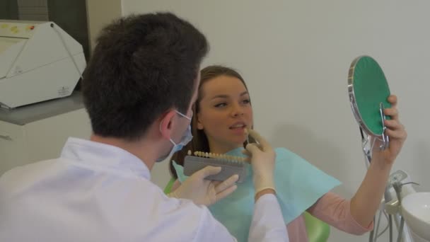 Uomo in laboratorio cappotto dentista cercando un nuovo colore dei denti per una giovane donna medico in maschera e la sua paziente donna sta guardando specchio sorridente clinica dentale — Video Stock