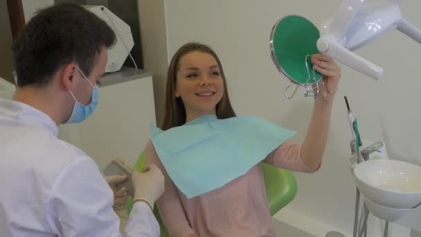 Zahnarzt probiert eine neue Zahnfarbe für eine Ärztin in Maske und Handschuhen aus und seine Patientin blickt lächelnd in den Spiegel einer sprechenden Zahnklinik — Stockvideo