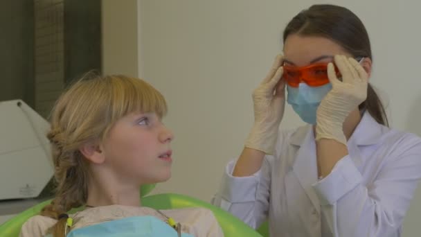 Dentista se pone una gafas en el uso de la lámpara dental UV Paciente niña Doctor en máscara está tratando un diente de un paciente en la sala de tratamiento dental Visita al dentista — Vídeo de stock