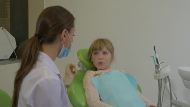 Dentysta rozmawia z dziewczyną uspokaja lekarz pacjenta w Mask bada zęby pacjenta wyjaśnia coś Klinika dentystyczna wizyta u dentysty — Wideo stockowe