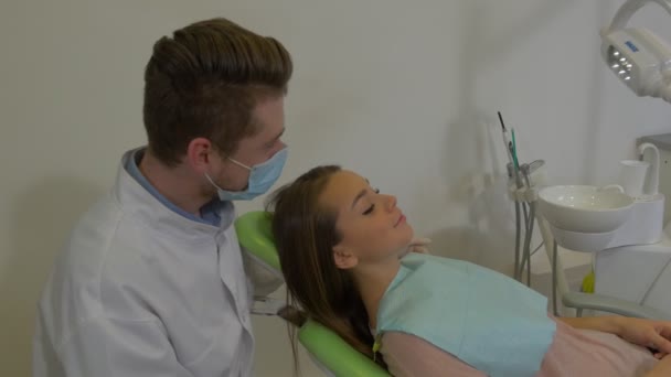 Ο οδοντίατρος κάθεται πίσω από το κεφάλι ενός πελάτη που μιλάει άνθρωπος με μάσκα είναι εξετάζοντας ένα δόντι μιας ασθενούς γυναίκα με χαρτοπετσέτα στο θώρακα της οδοντιατρικής κλινική επίσκεψη — Αρχείο Βίντεο