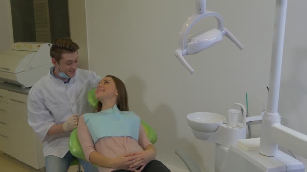 Tandläkare pratar med en kvinna vänlig lycklig klient ler liggande på en stol ung blond läkare och en patient tandklinik undersökning av en tänder — Stockvideo