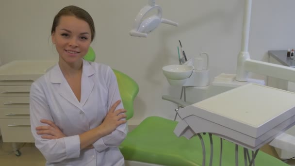 Jeune femme dentiste est assis sur la chaise Souriante fille avec ses bras croisés regardant la caméra Jeune médecin habillé en blouse de laboratoire à la clinique dentaire — Video