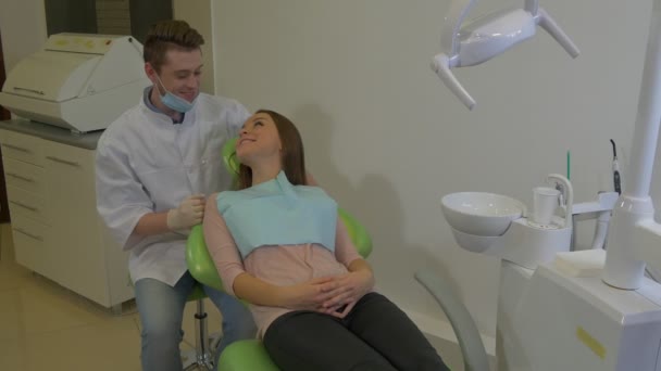 Tandarts praat met een klantvriendelijke jonge vrouw glimlacht op een stoel jonge blonde arts en een patiënt tandheelkundige kliniek onderzoek van een tanden — Stockvideo